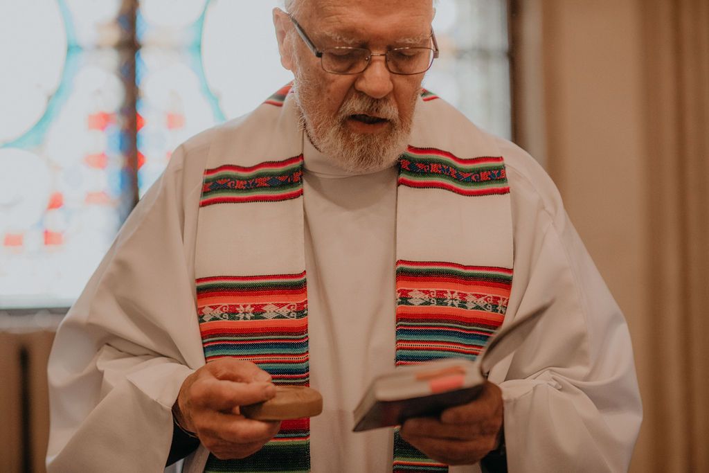Elderly priest in white robes and colorful stole reads from a small book during a religious ceremony.