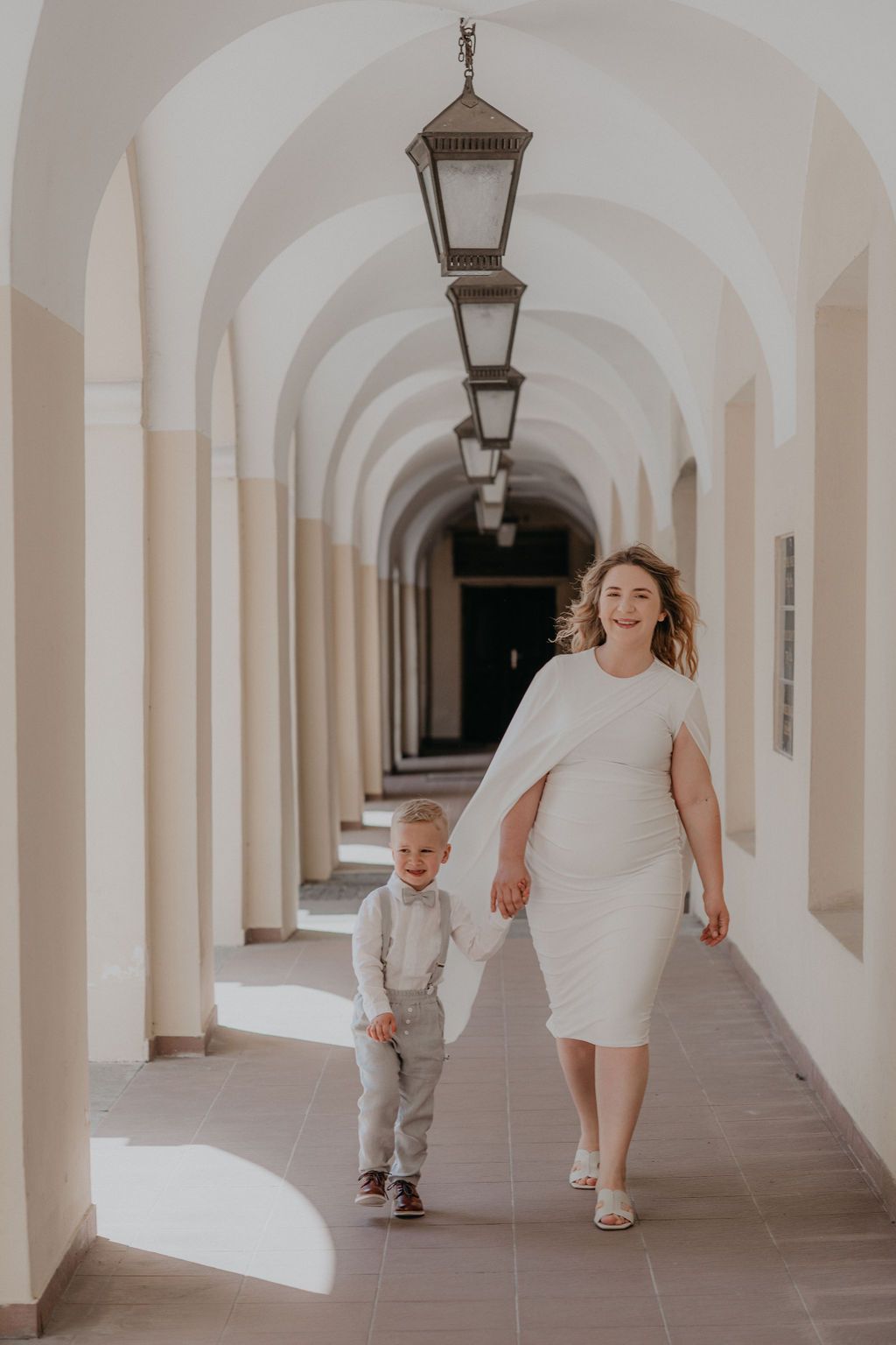 Woman in white dress walks with young boy in suspenders through elegant arched corridor with hanging lanterns.
