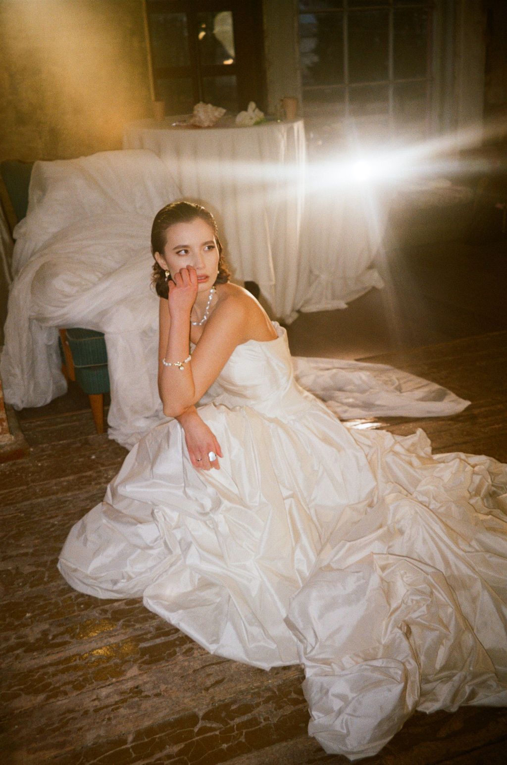 Bride in white ball gown sitting on wooden floor, illuminated by bright light, wearing gold jewelry in rustic setting.
