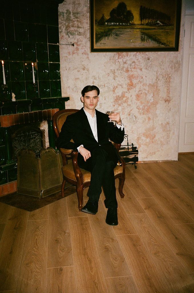 Young man in black suit sitting in vintage chair holding a drink, beside a fireplace with rustic walls.
