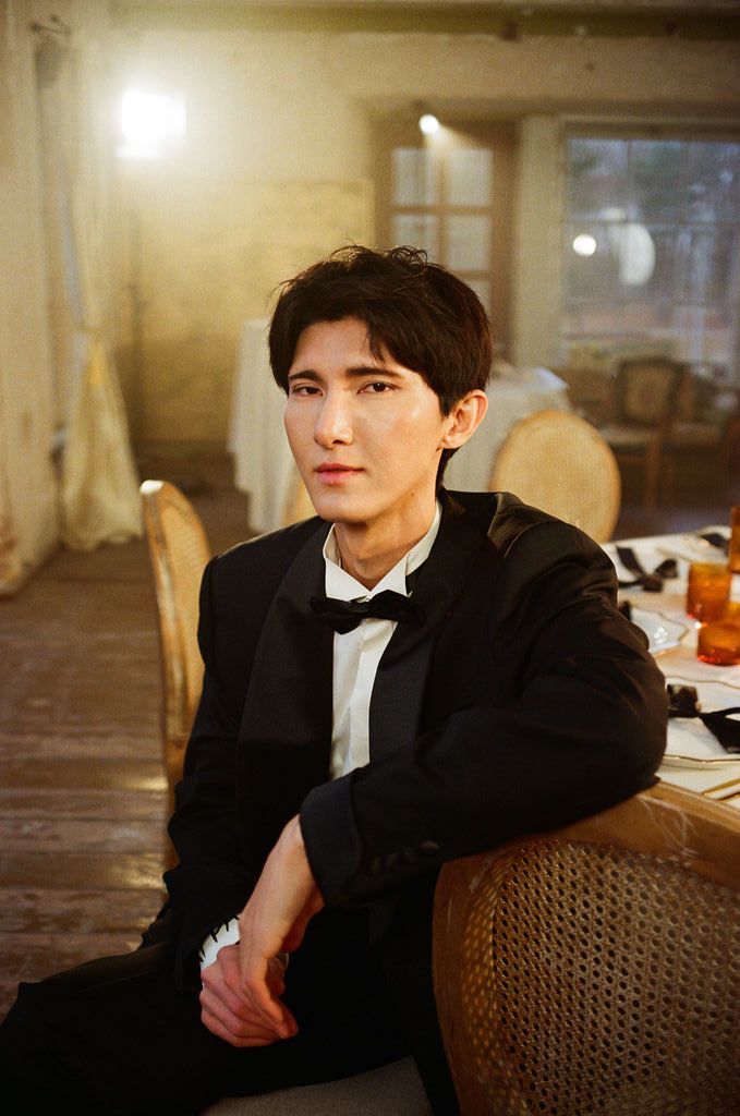 Young man in black tuxedo with bow tie seated in elegant room with warm lighting.