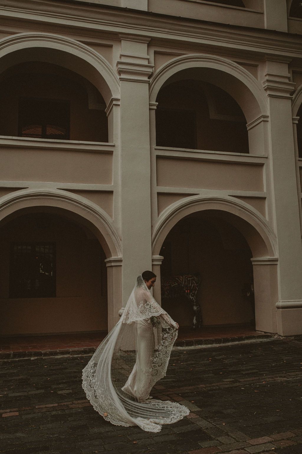 Bride in lace wedding dress and long veil standing before arched colonial building facade.