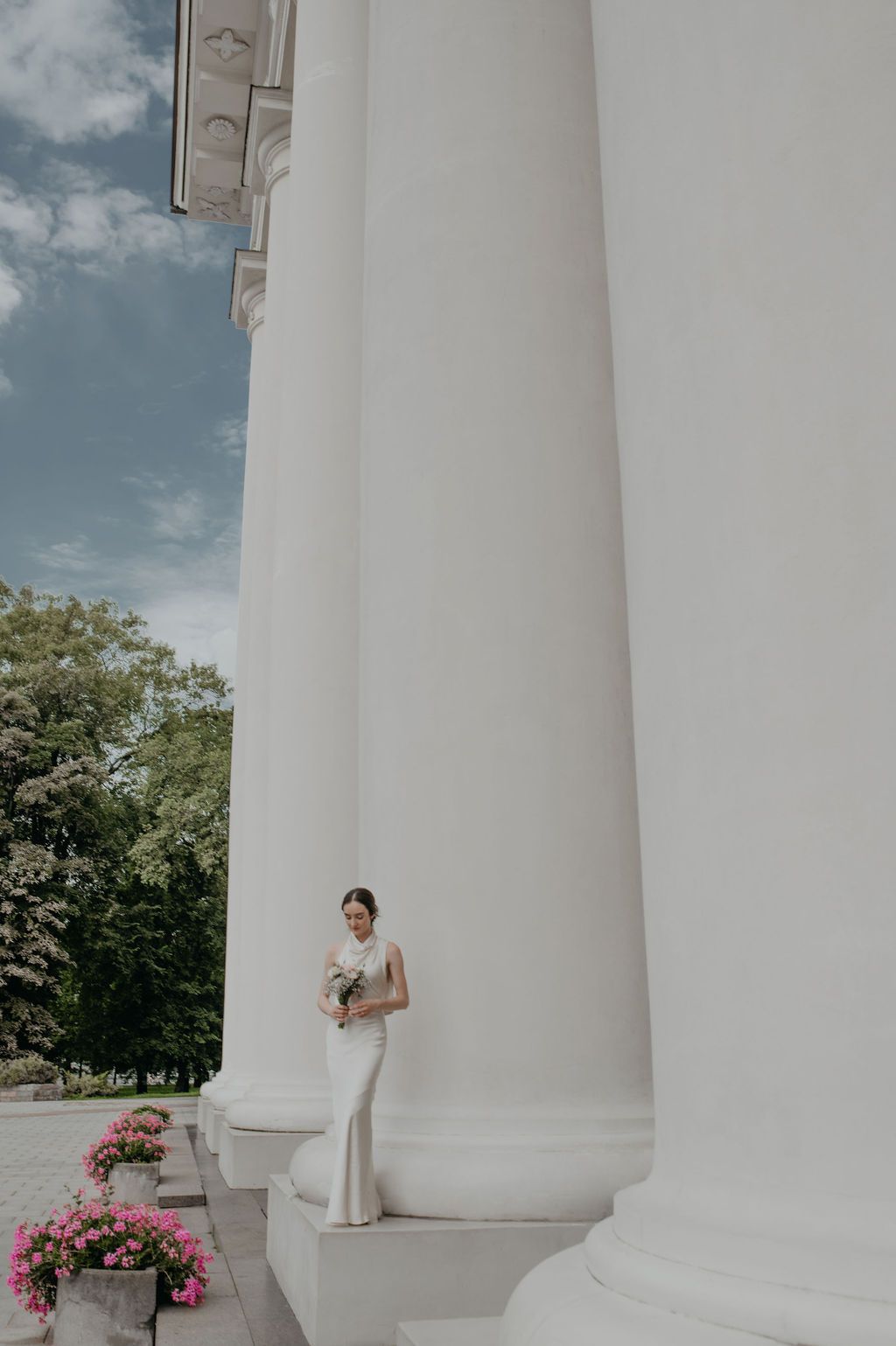 Bride in white gown holding bouquet stands between large white columns of elegant building on sunny day.