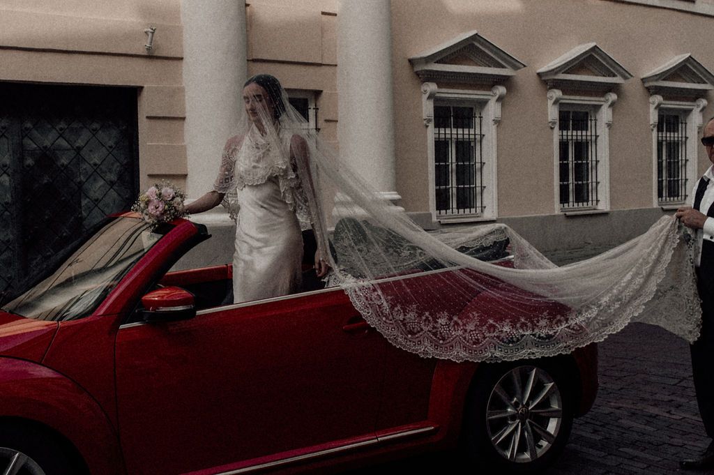 Bride in white wedding dress with flowing lace veil stands beside a red convertible car.