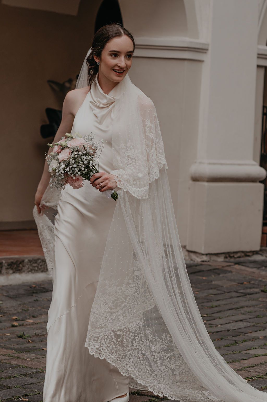 Elegant bride in a white satin halter gown with lace veil holding a pink bouquet outdoors.