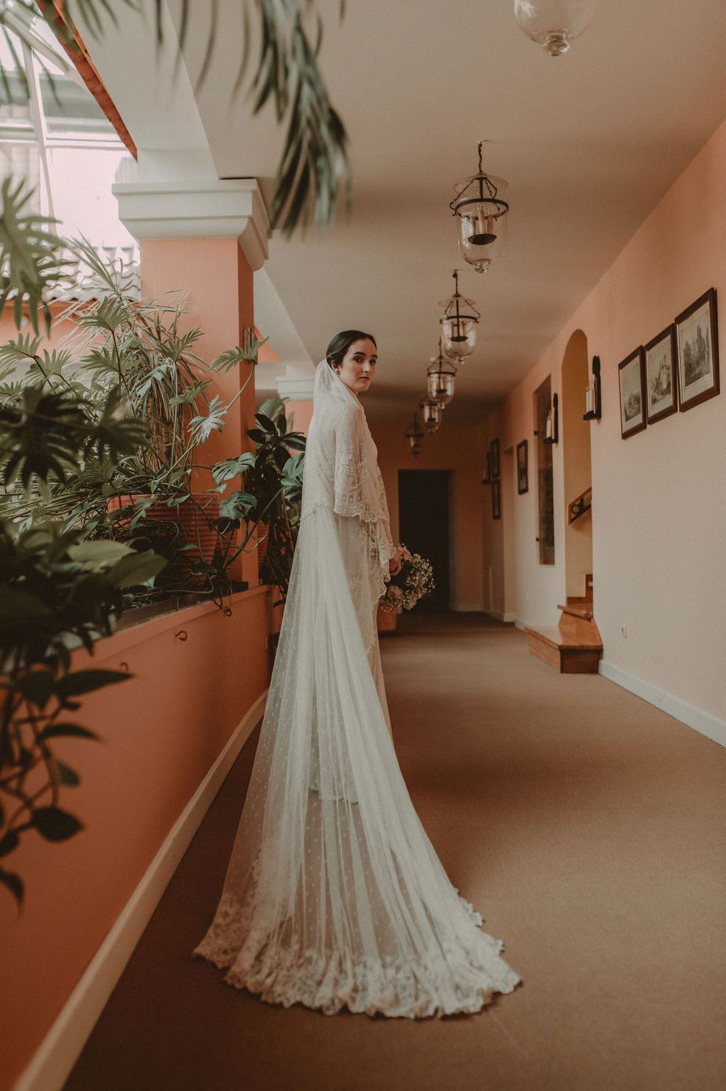 Elegant bride in lace wedding dress with cathedral veil posing in warmly lit corridor.