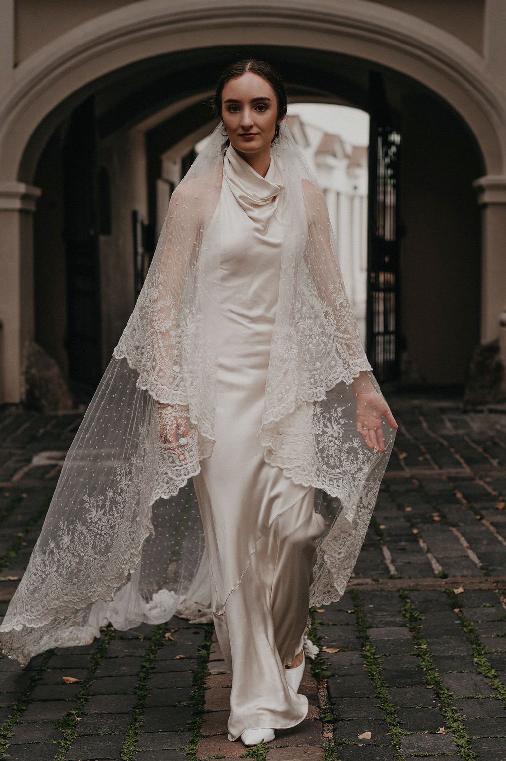 Bride in silk wedding gown with lace veil draped over shoulders walks through stone archway on cobblestone path.