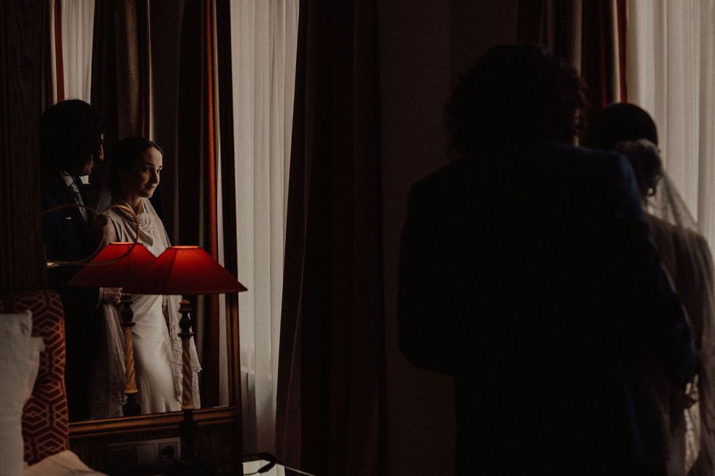 Bride in white dress reflected in mirror while being helped by attendants in dim room.