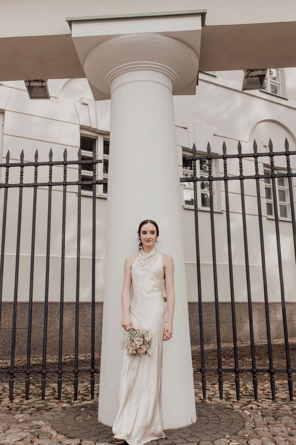 Bride in elegant white gown holding bouquet stands before white column and iron fence at classical building.