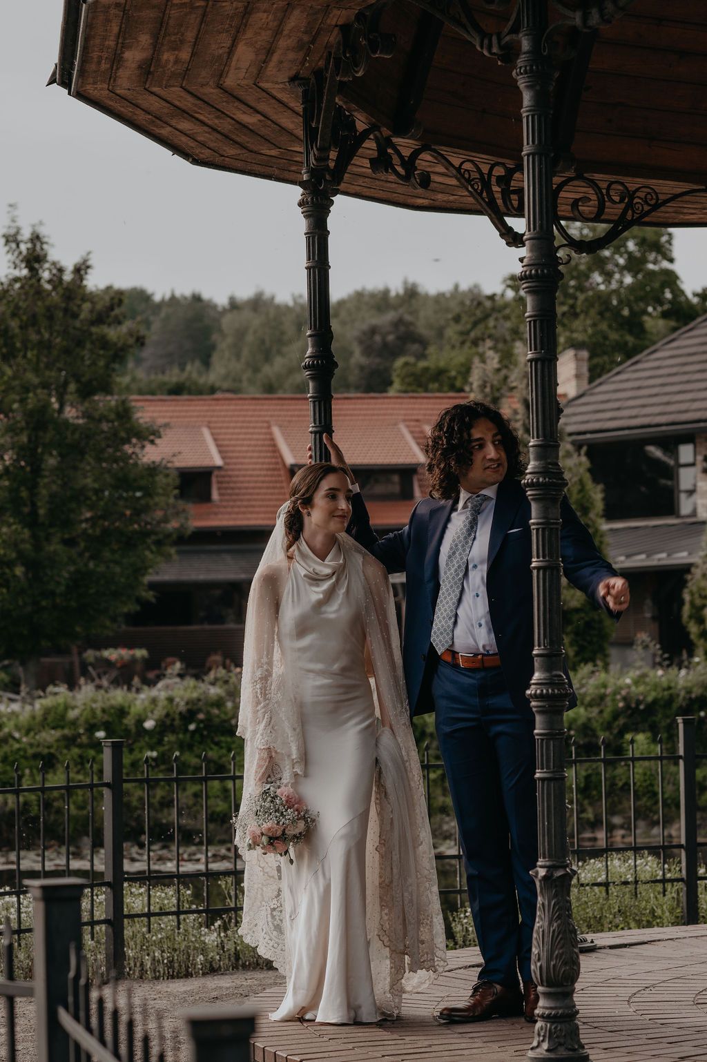 Bride in white gown with lace cape and groom in navy suit stand under ornate gazebo at outdoor wedding venue.