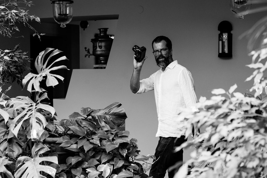 Black and white photo of a bearded man in white shirt holding a camera among tropical monstera plants outdoors.