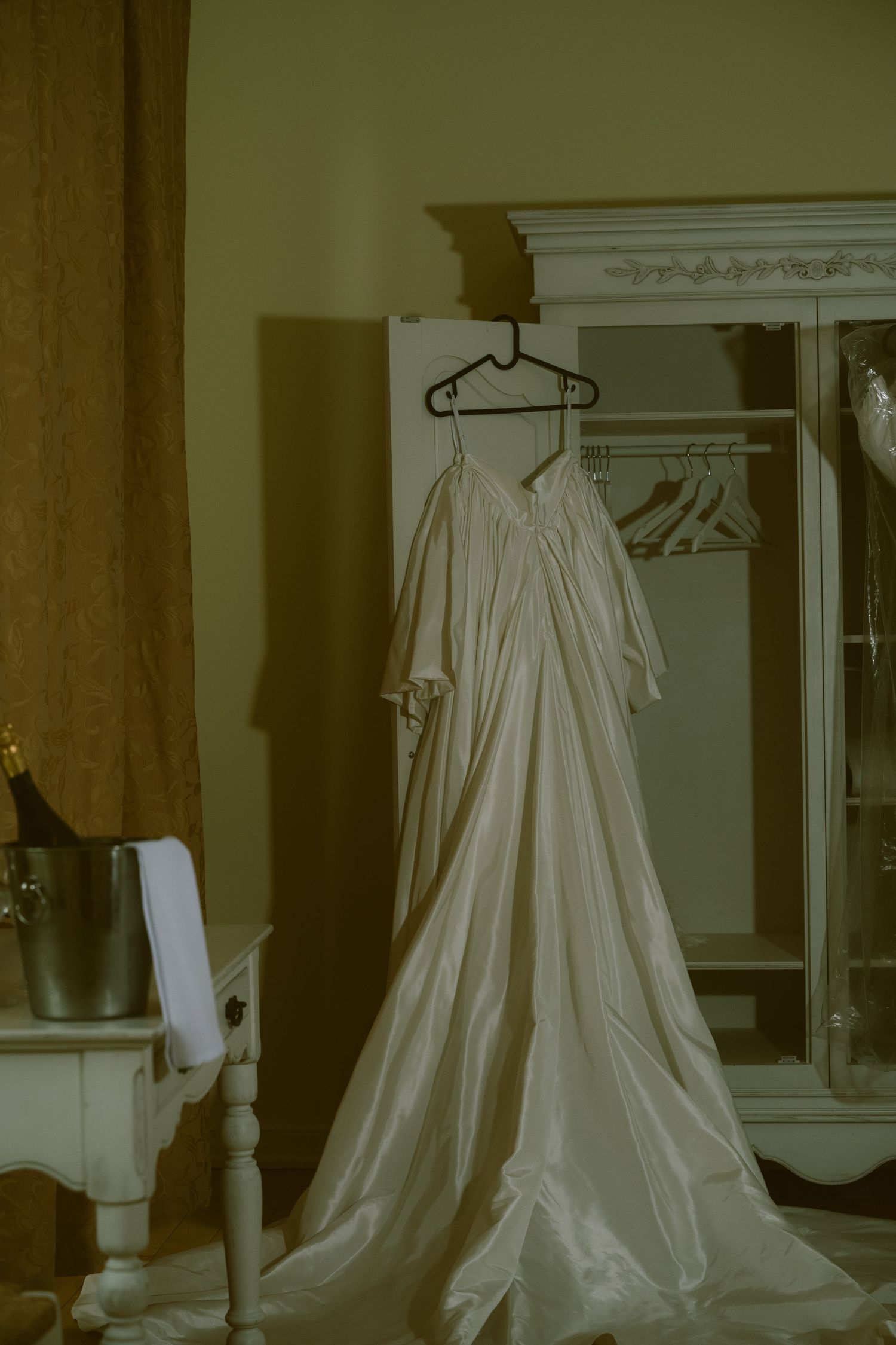 Elegant white wedding dress hanging on a hanger in front of an ornate white armoire in a softly lit room.