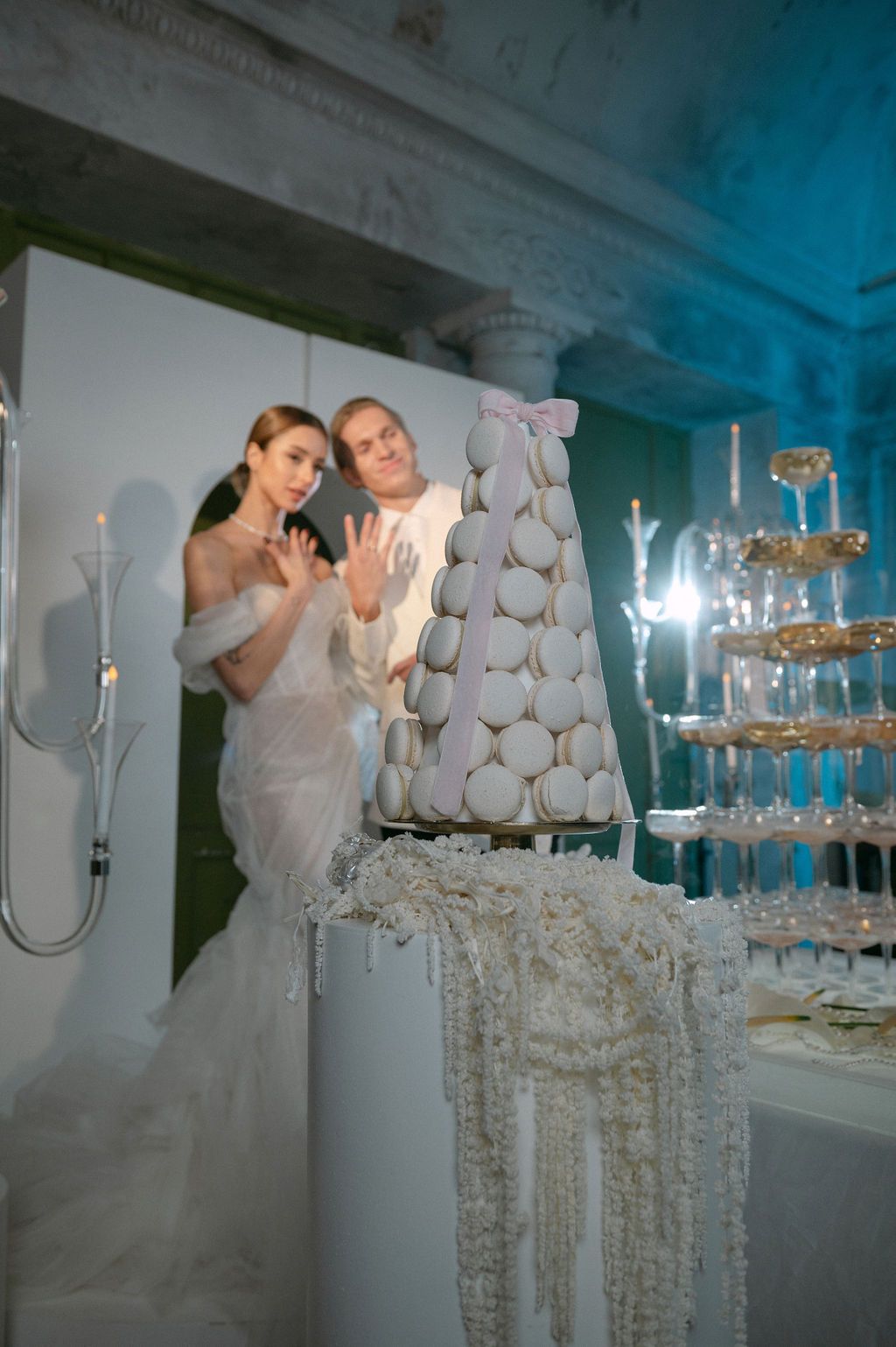 Bride and groom posing behind elegant macaron tower and champagne glass pyramid at luxury wedding reception.