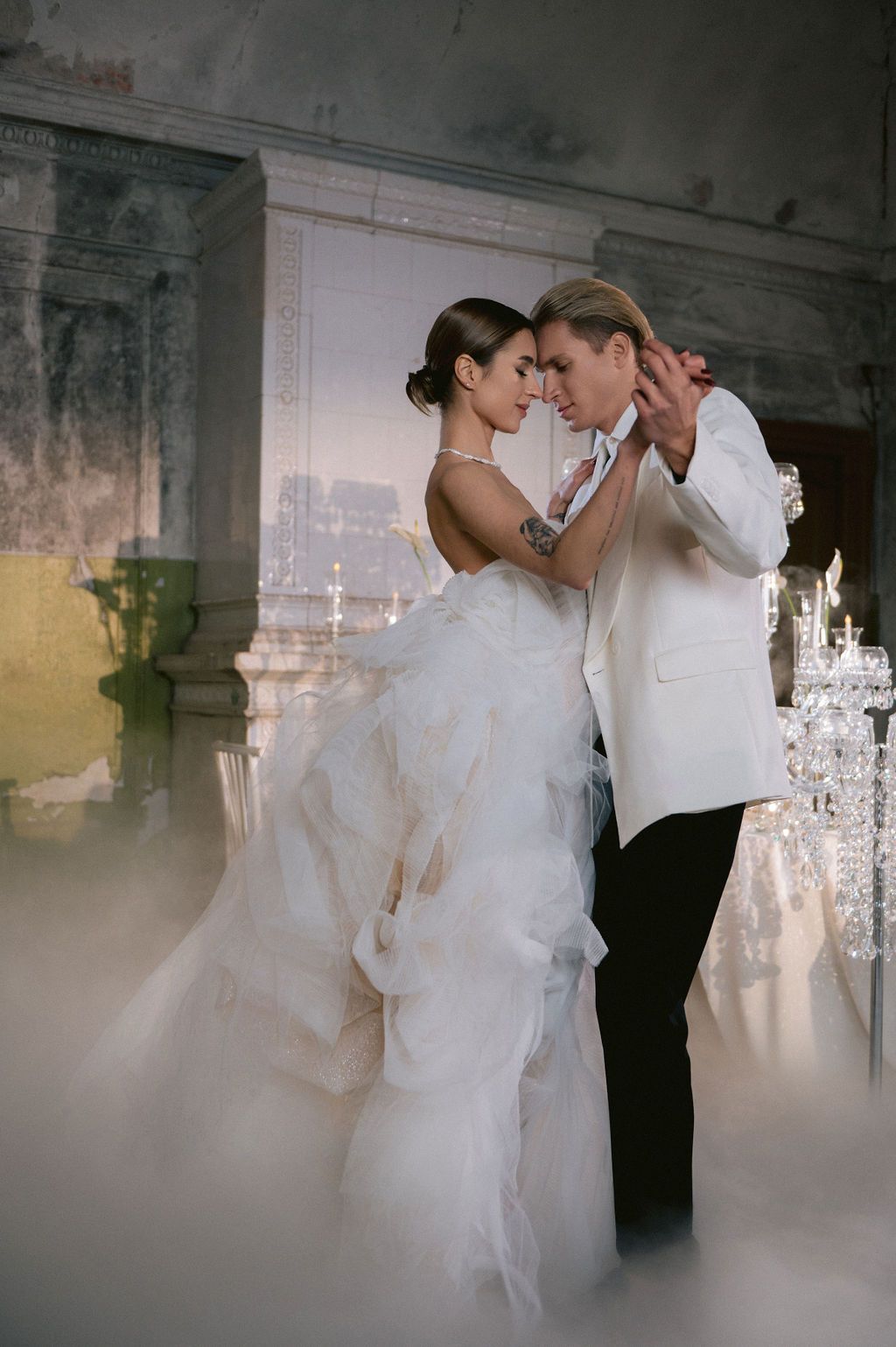 Bride and groom share first dance in elegant ballroom, bride in ruffled white gown, groom in white tuxedo jacket.