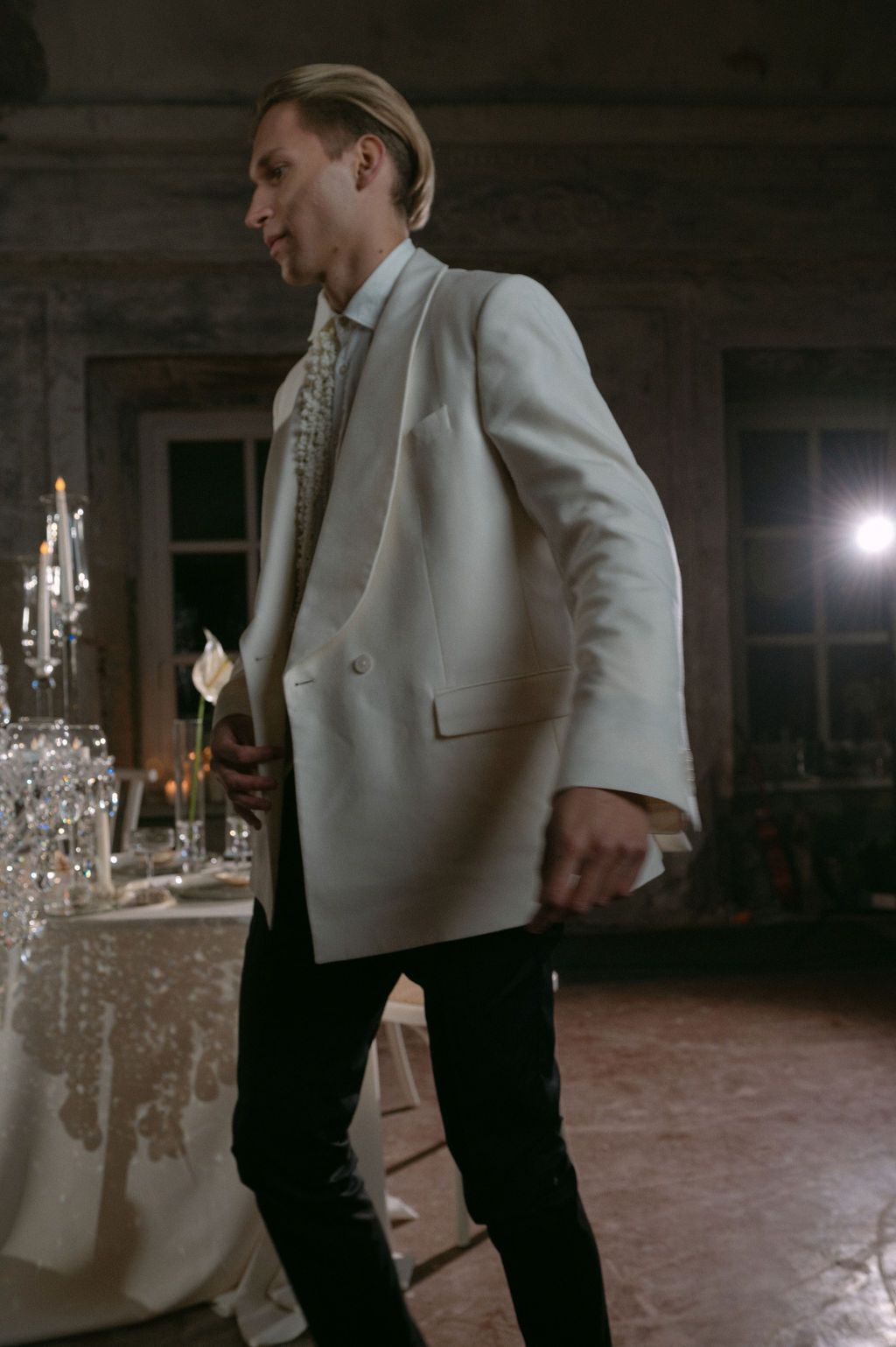 Groom in white tuxedo jacket with black trousers holding a calla lily in a dimly lit elegant reception hall.