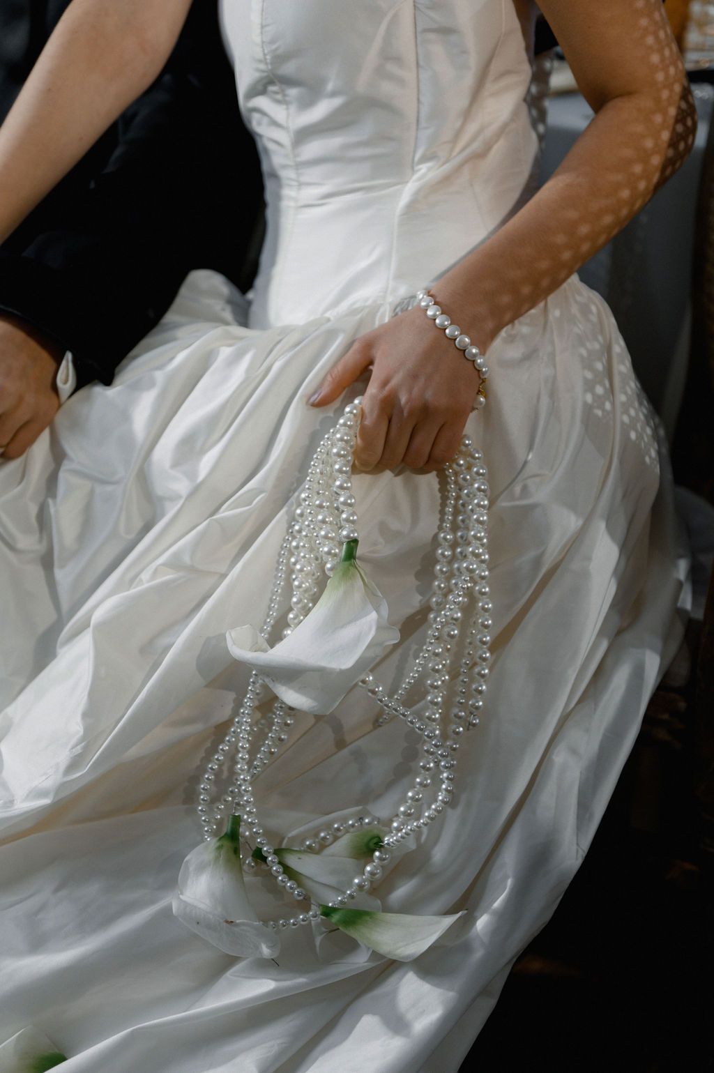 Bride in white satin gown holding pearl necklaces and white calla lily bouquet with green accents.
