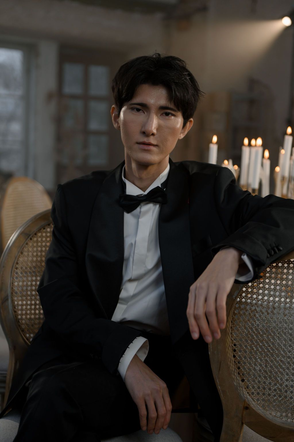 Elegant man in black tuxedo and bow tie seated in ornate chair with candles glowing in background.