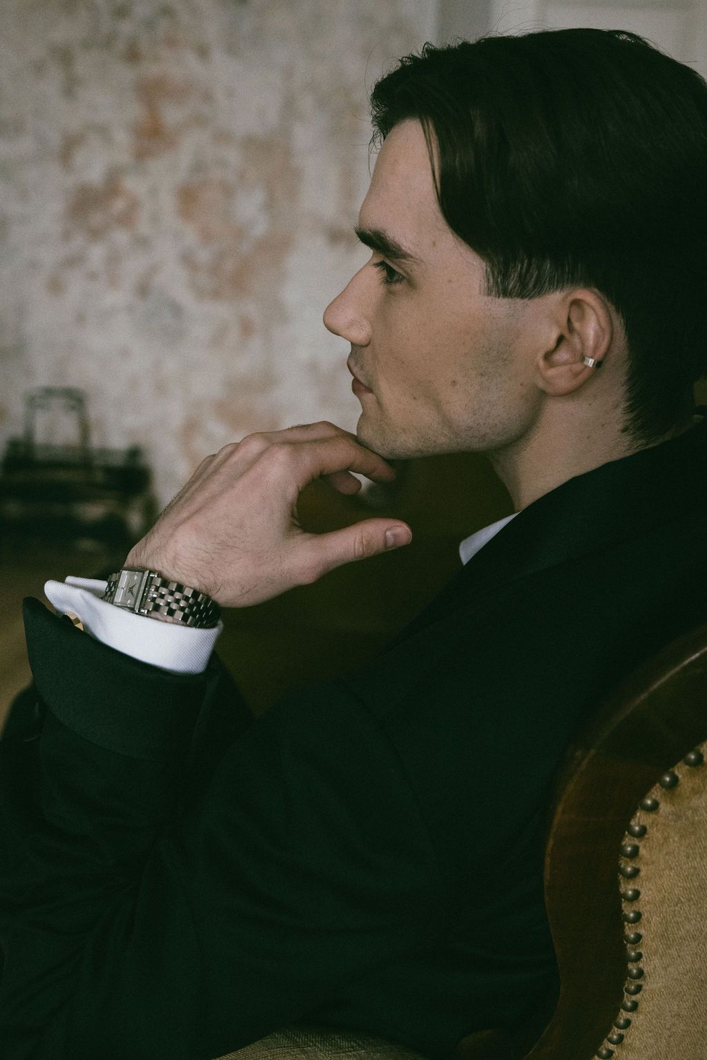 Sophisticated man in black suit seated in profile view with hand on chin in vintage interior.