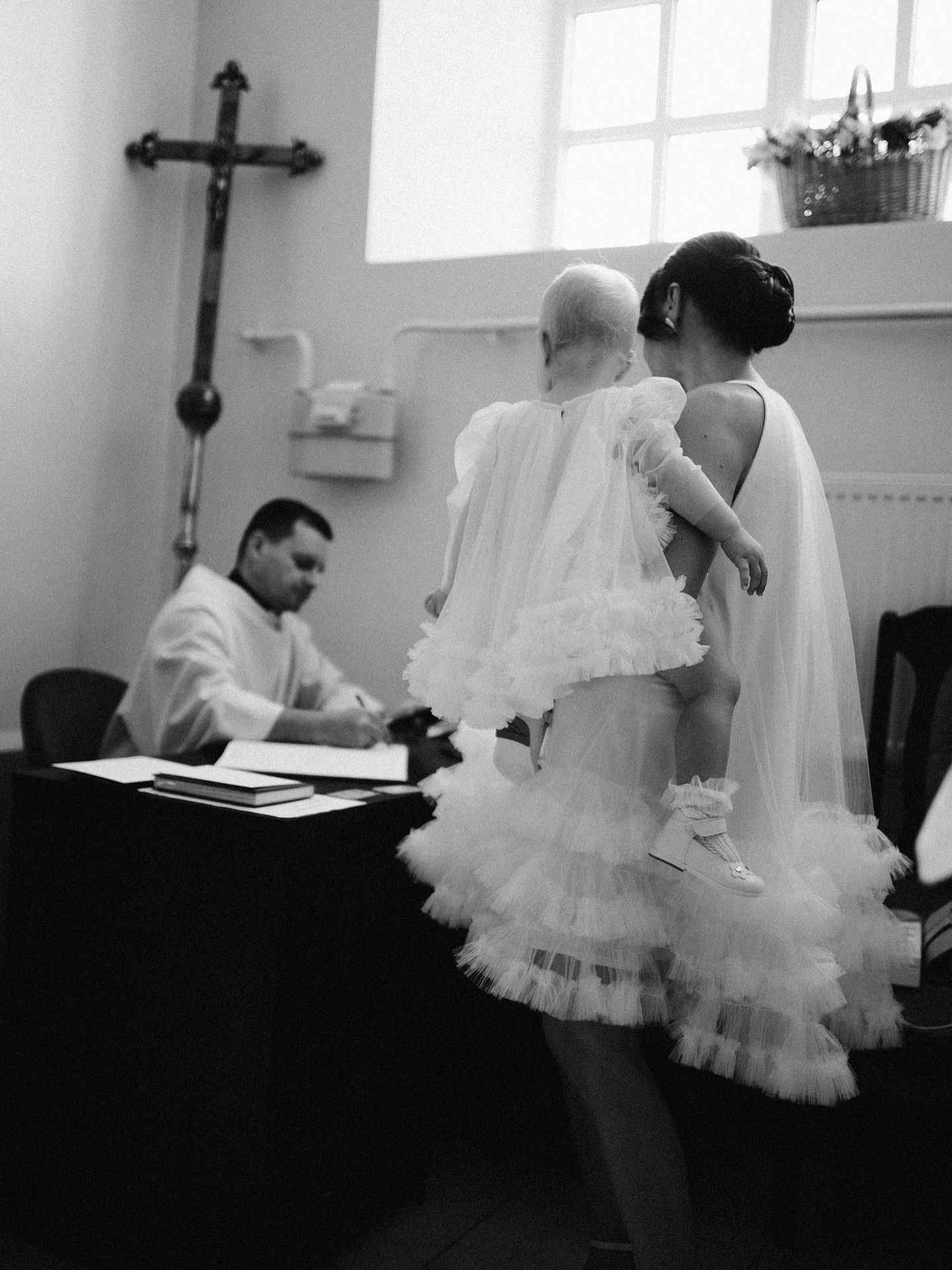 Black and white photo of a bride holding a baby in white dress before a priest at a church baptism ceremony.