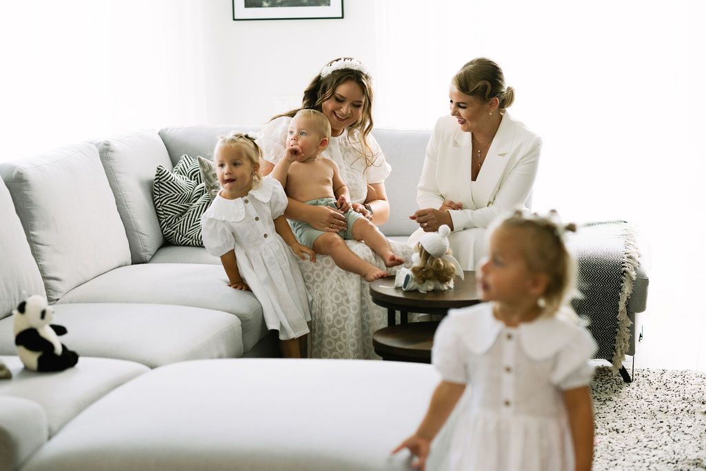 Two women in white outfits sit on a gray sofa with three young children in a bright living room.