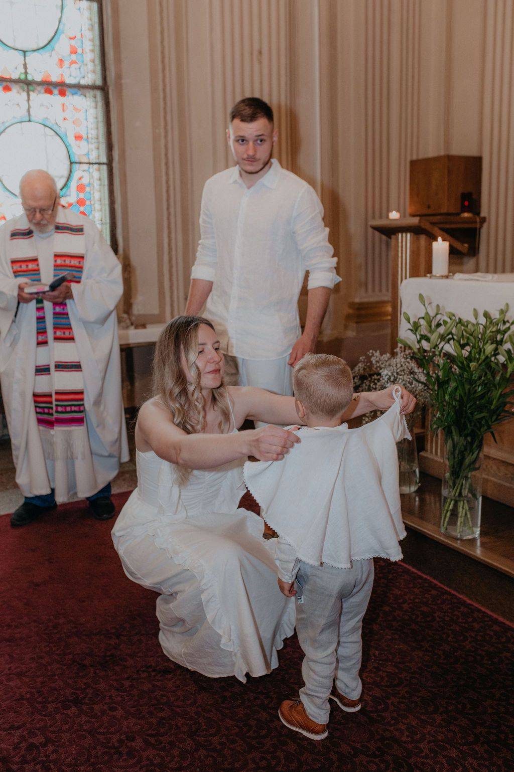 A mother in white dress helps her young son with his outfit during a church baptism ceremony with a priest in the background.