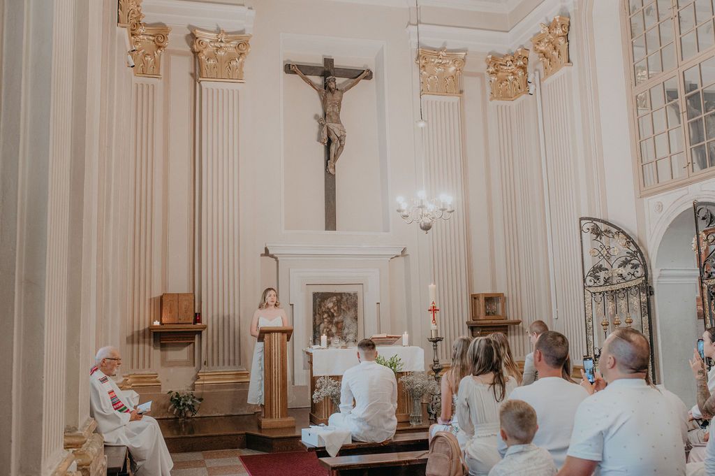 A woman speaks at a podium during a Catholic church ceremony with a crucifix and white columns in the background.