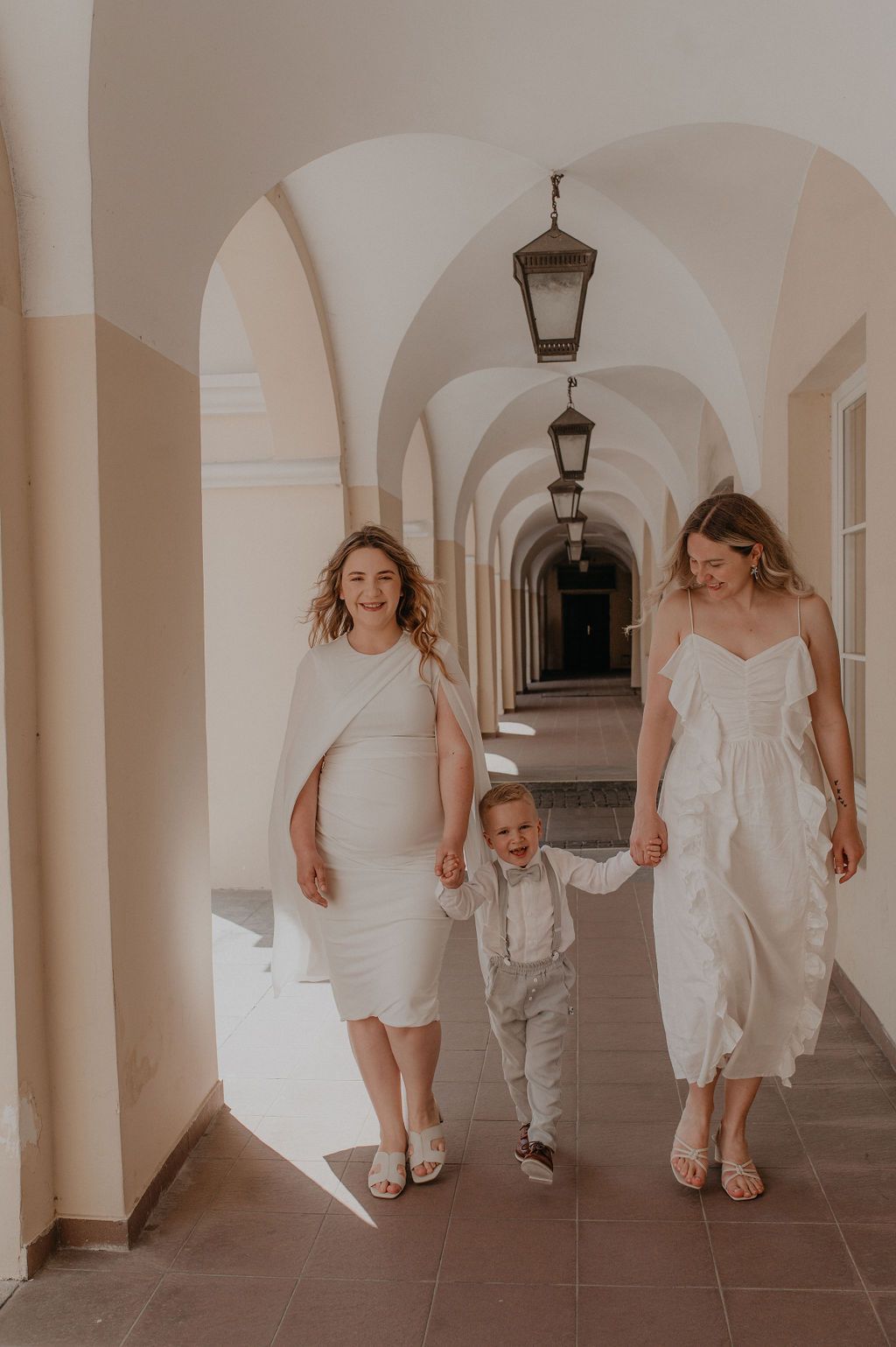 Two women in white dresses walk with a young boy in a suit through an elegant arched corridor.