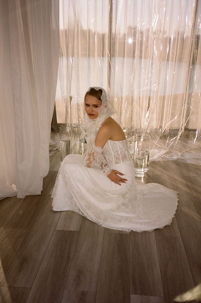 Bride in white lace wedding gown and veil sits gracefully on hardwood floor beside sheer curtains.