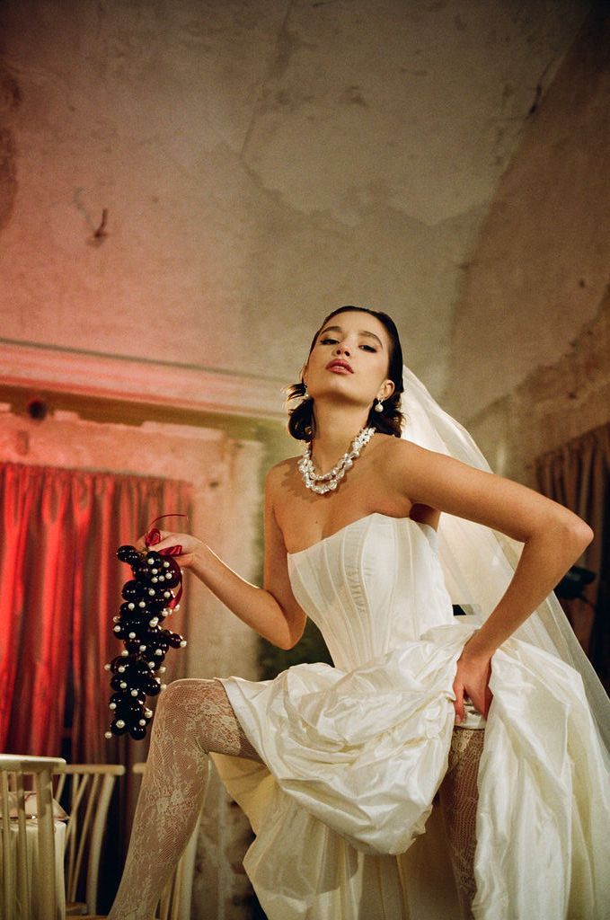 Woman in white strapless corset wedding gown with veil and pearl jewelry poses elegantly in vintage setting.