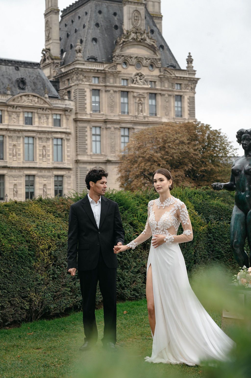 Elegant couple in wedding attire posing in Tuileries Garden with the Louvre Palace in Paris background.