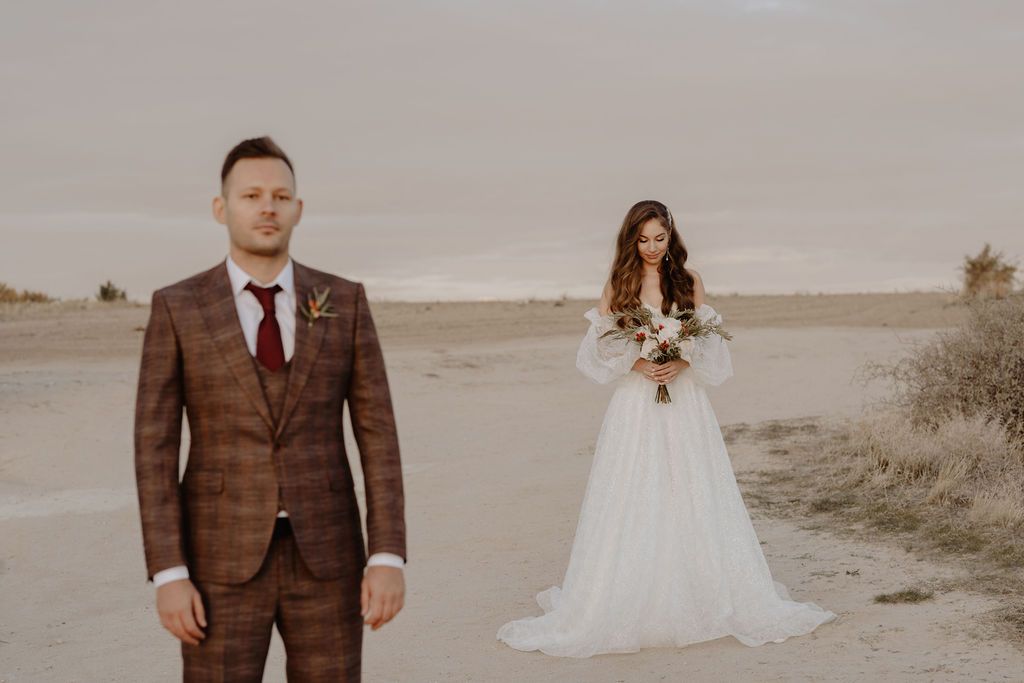 A bride in a white gown and groom in a brown plaid suit pose in a sandy desert landscape during golden hour.