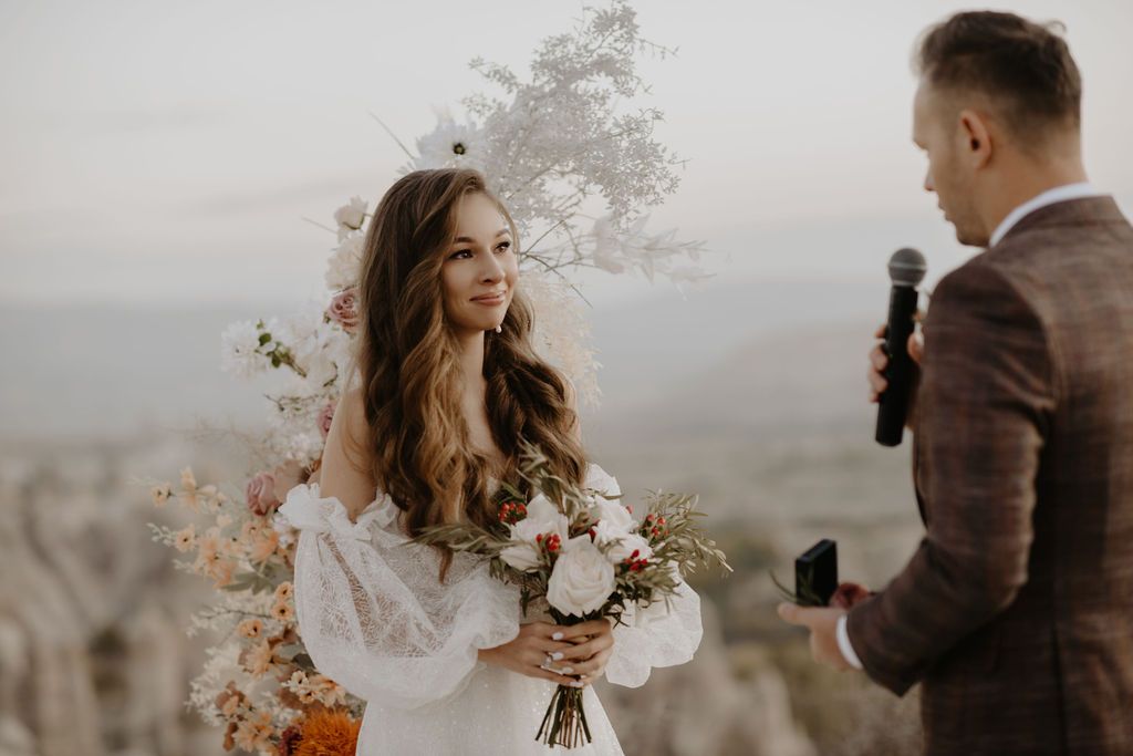 Bride smiling at groom during outdoor wedding ceremony with floral bouquet at scenic overlook.