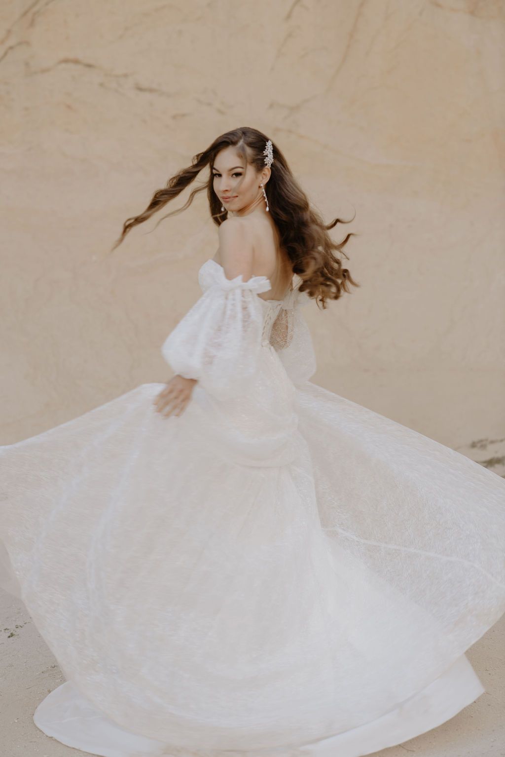 Bride twirling in an elegant off-shoulder white ball gown with puff sleeves against a sandy backdrop.