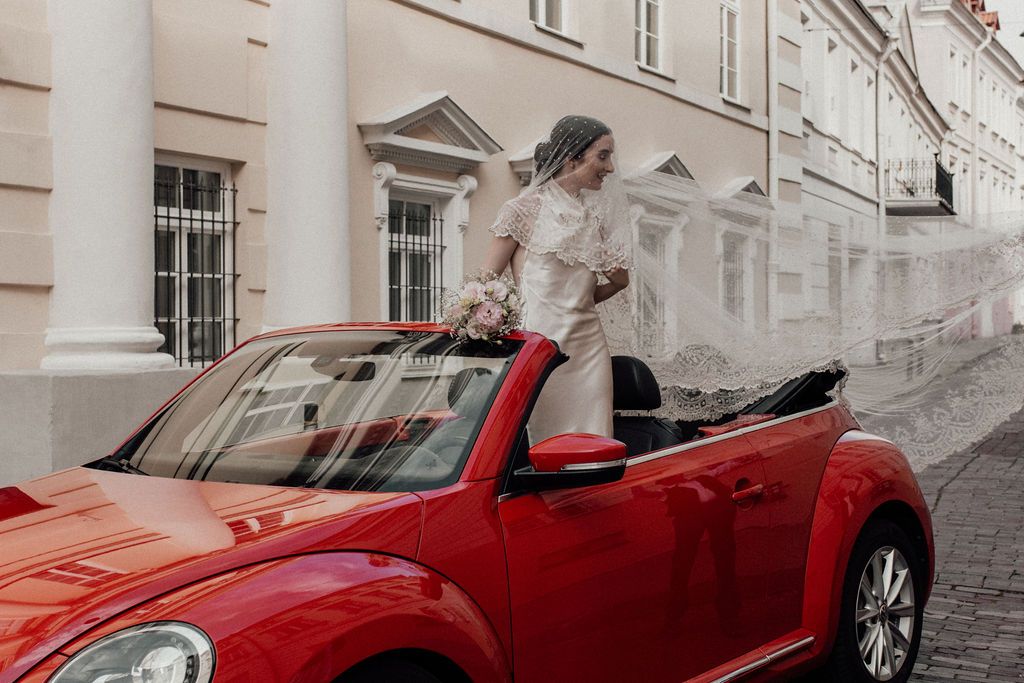 Bride in white dress and veil standing in red convertible VW Beetle on a cobblestone street.