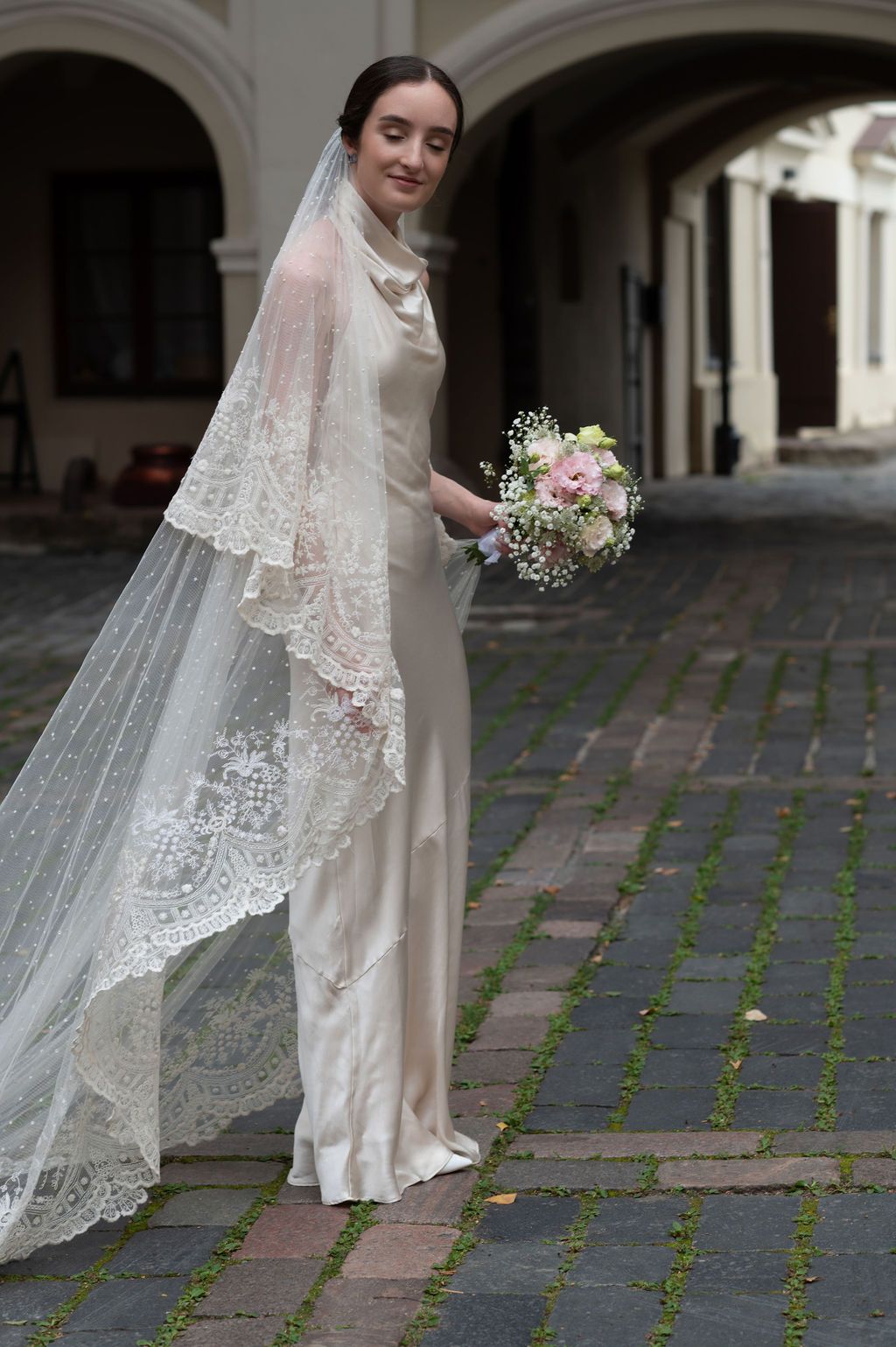 Bride in ivory satin gown with lace veil holding pink bouquet on cobblestone courtyard.