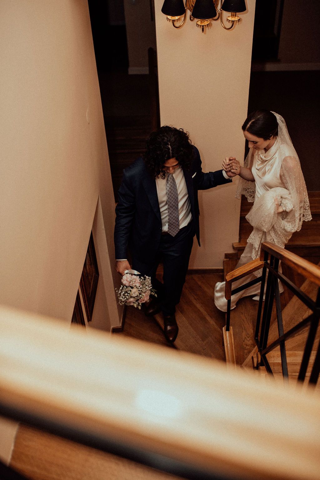 Aerial view of groom guiding bride down indoor staircase, holding hands, with bridal bouquet in hand.