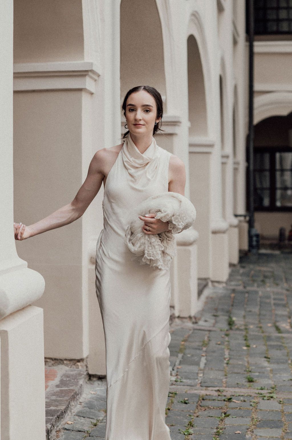 Bride in ivory satin halter gown holding a fluffy bouquet, posing by white columns in an elegant courtyard.