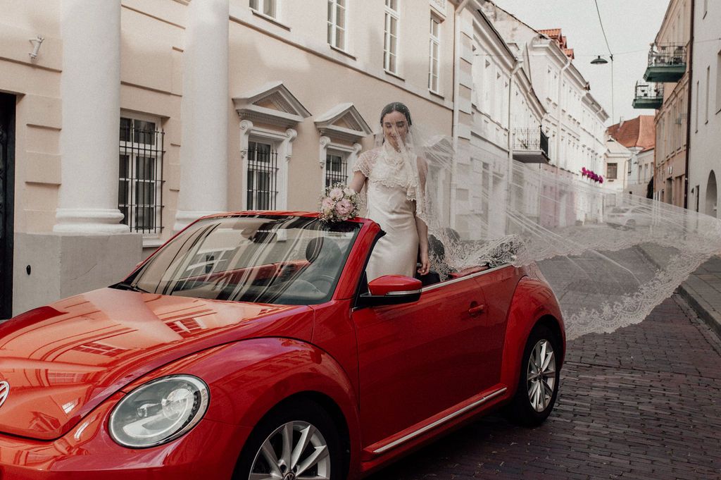 Bride in white dress standing in red VW Beetle convertible on cobblestone European street with flowing veil.