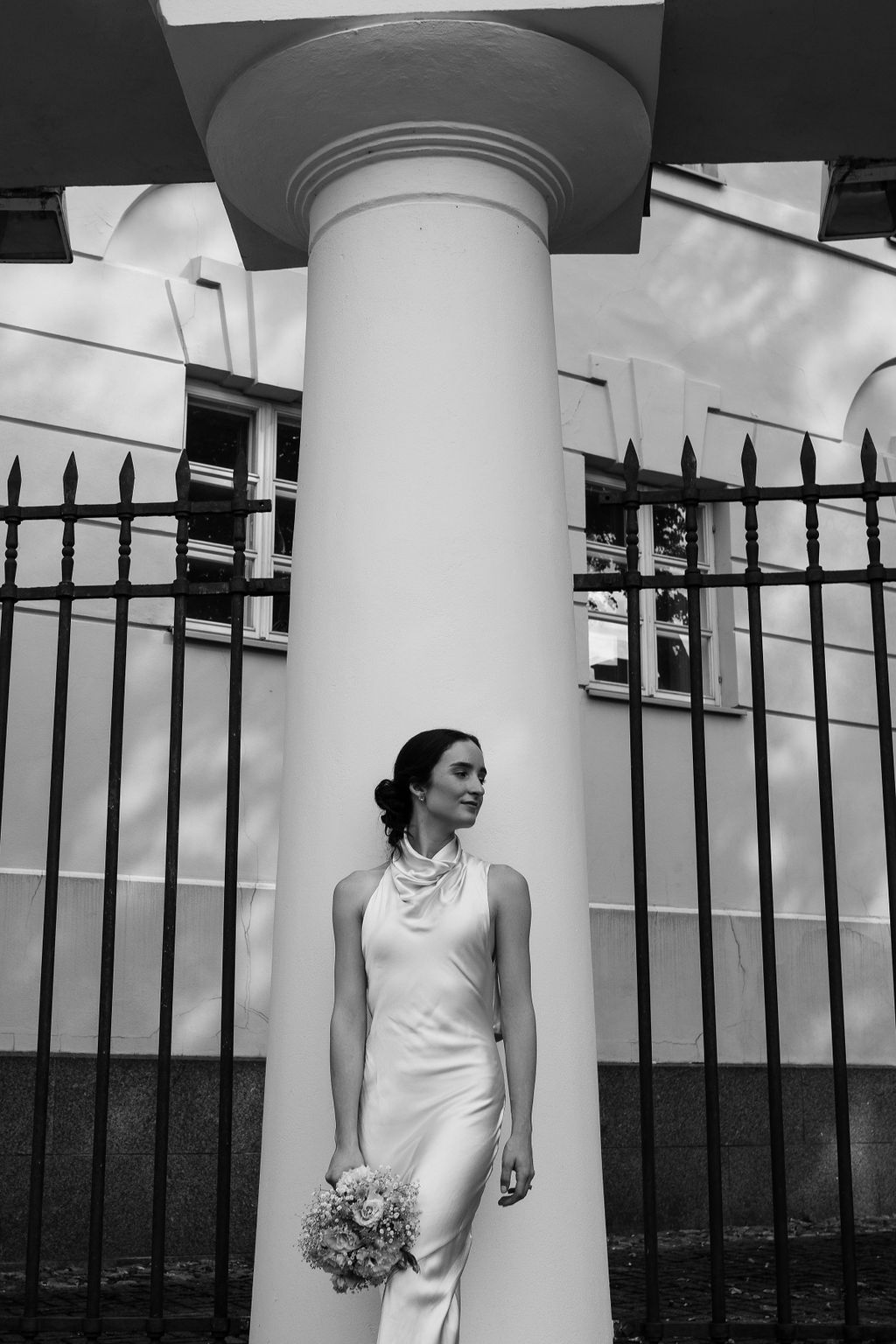 Elegant bride in a sleek halter gown holds a bouquet beside a white column and iron fence in a black and white portrait.