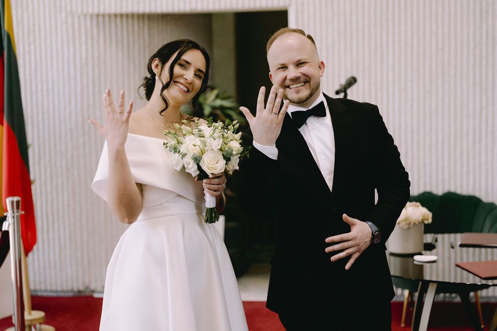 Newlywed couple showing wedding rings, bride in white dress holding bouquet, groom in black tuxedo, red carpet ceremony.