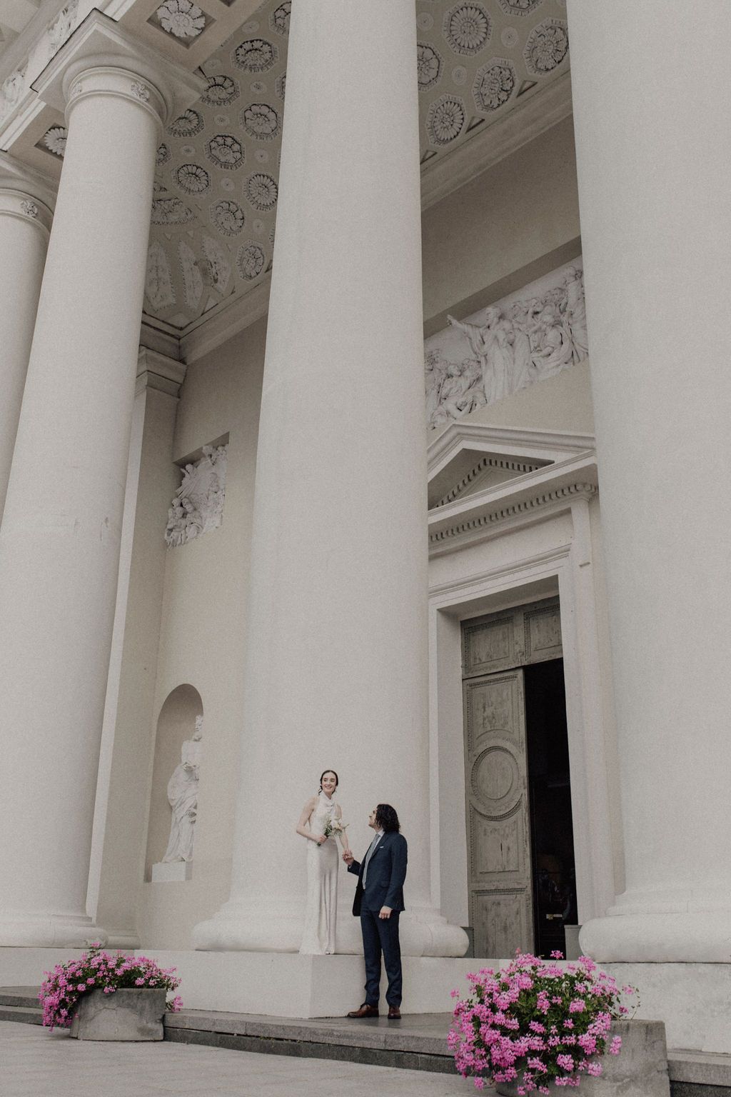 Bride and groom stand before grand white neoclassical building with tall columns and ornate relief sculptures.