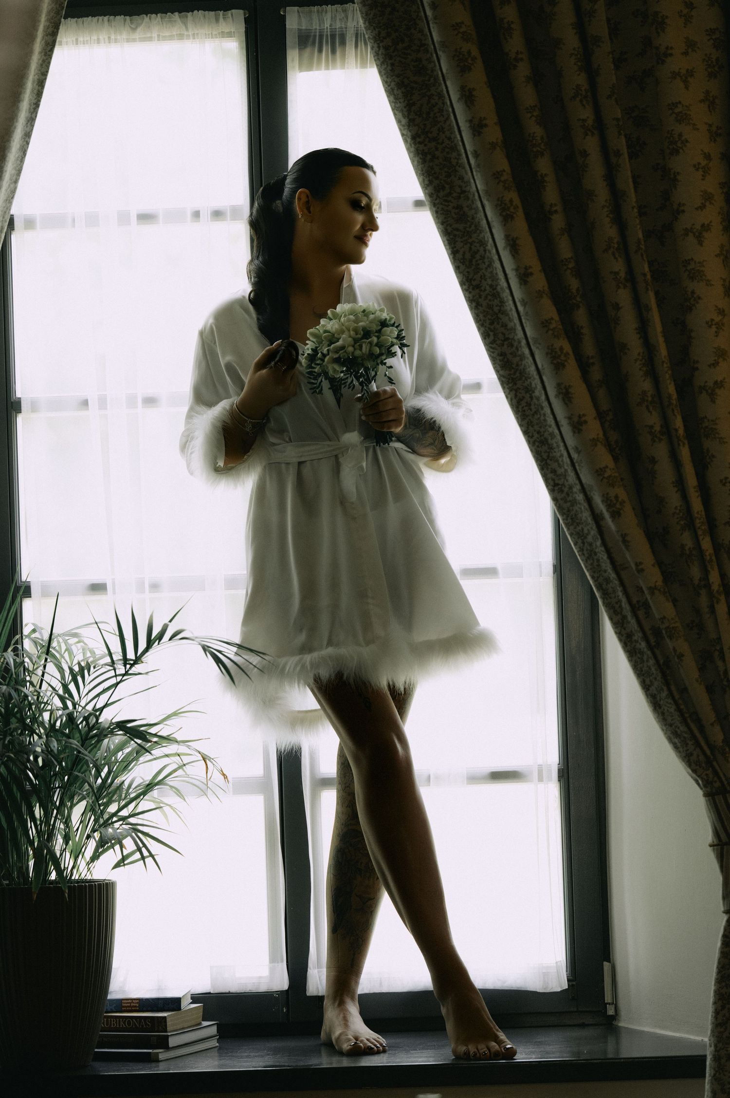 Bride in a white satin robe with feather trim stands barefoot on a windowsill holding a bouquet in soft natural light.