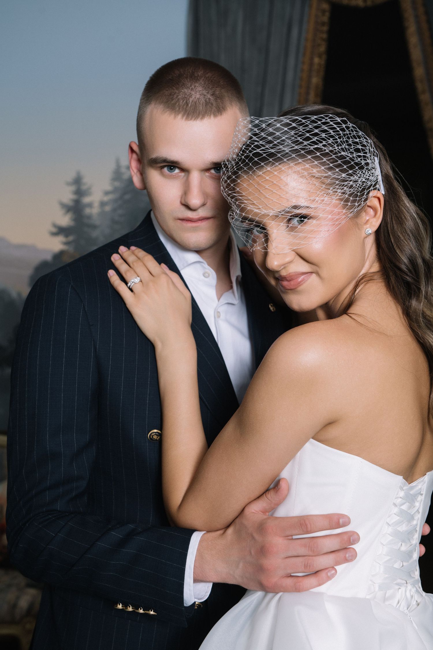 A smiling bride in a strapless white gown and birdcage veil embraces a groom in a navy pinstripe suit in an elegant, softly lit setting.