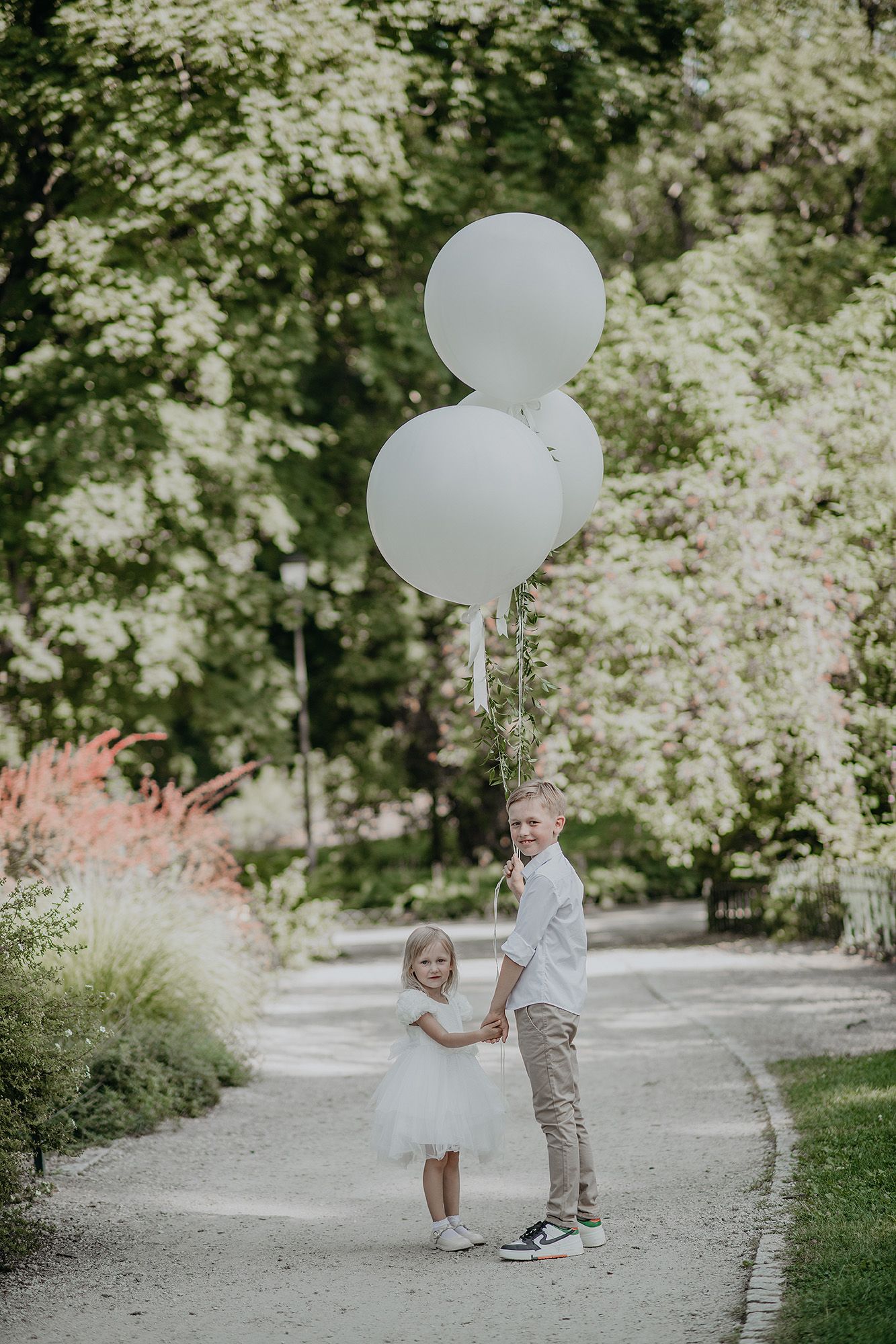 Two young children in white outfits holding white balloons on a tree-lined path.