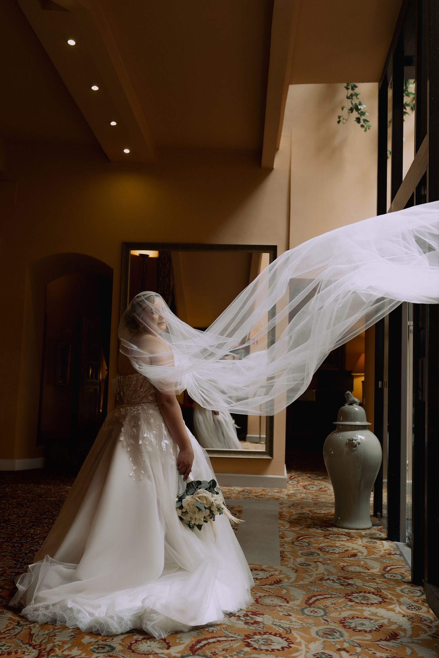 A flowing white wedding veil catches the wind dramatically in an elegant hotel hallway with patterned carpet.
