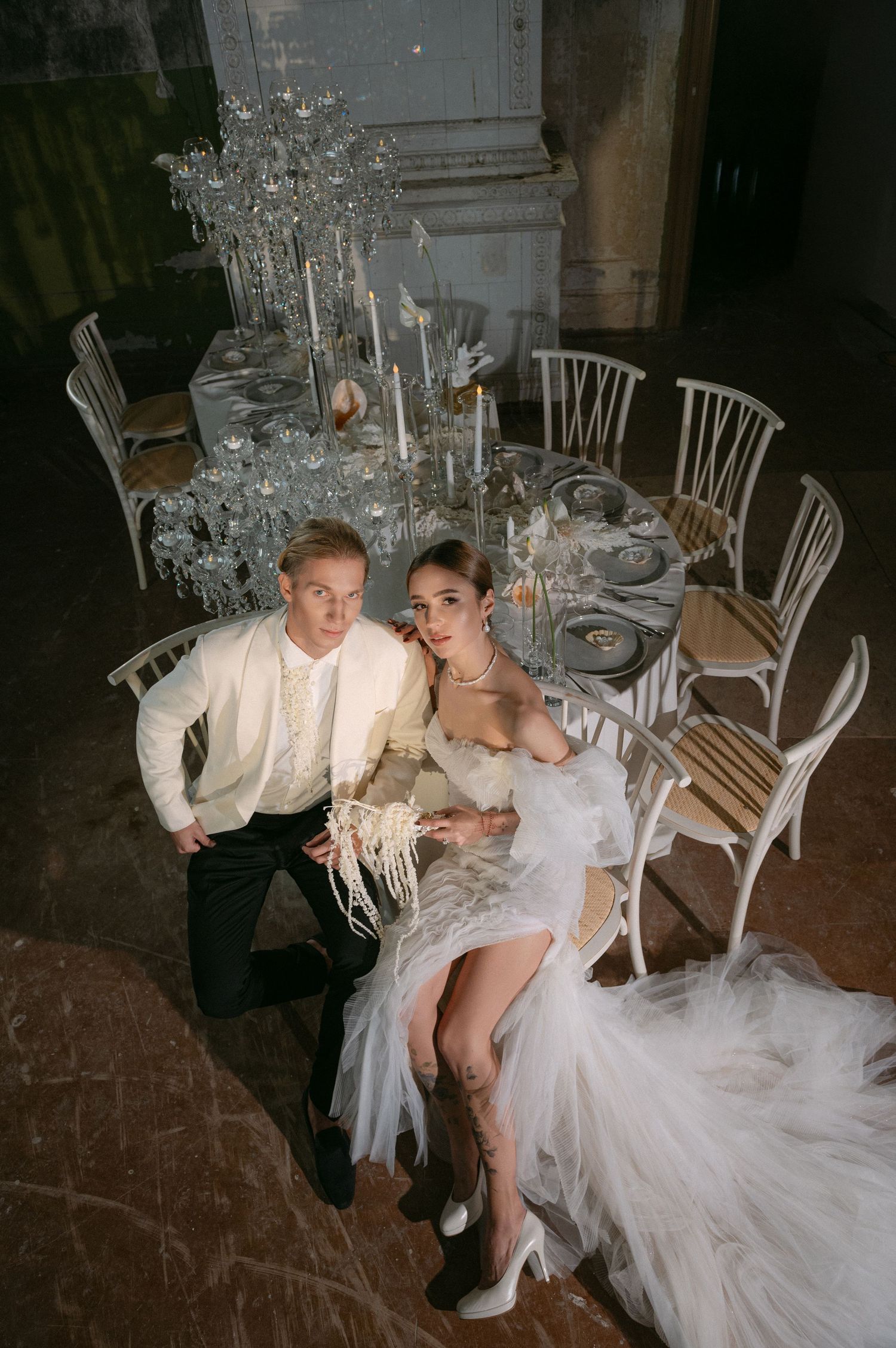 A couple in wedding attire shares an intimate moment while seated together on white chairs at their reception venue.