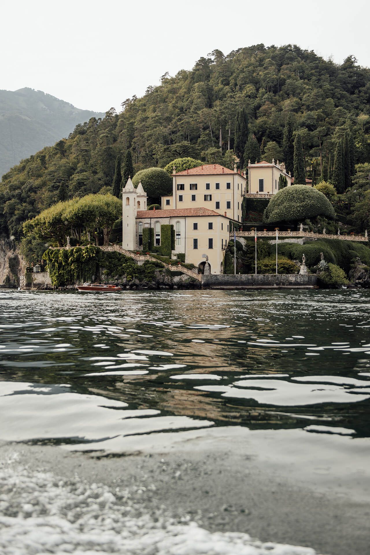 Prie Italijos ežero pakrantės driekiasi vilos ir pastatai, o fone matyti kalnų panorama.