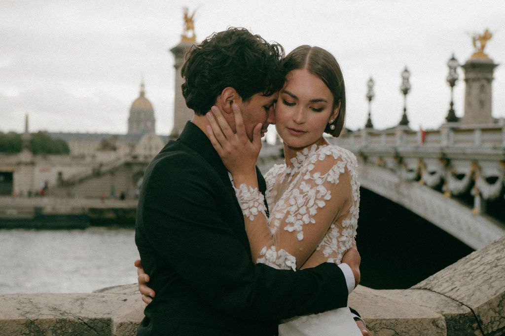Romantic couple embracing near Pont Alexandre III bridge in Paris, bride in lace gown and groom in black suit.