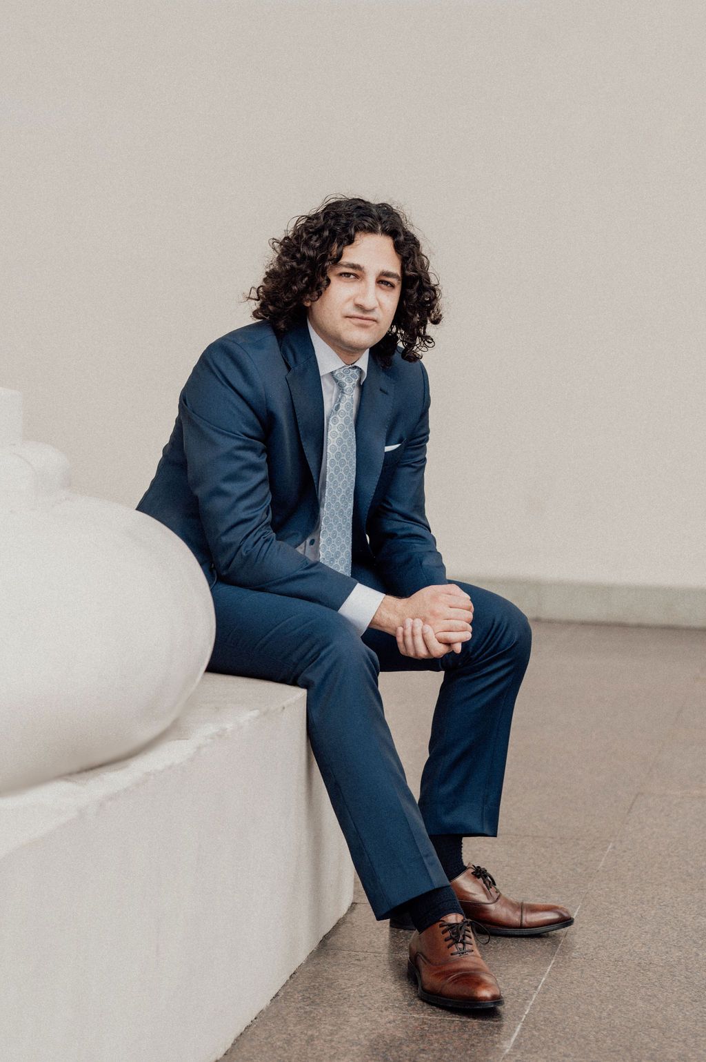 Young man with curly hair in navy blue suit and brown dress shoes sitting on white stone bench.