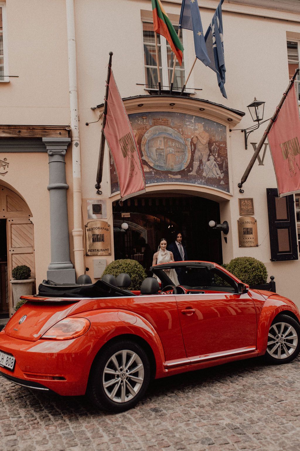 Newlyweds pose beside a red VW Beetle convertible outside an elegant building adorned with flags.